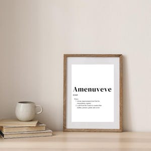 以下が含まれることがあります： 「Amenuveve」という単語が黒いフォントで書かれたフレーム入りのプリント。その下に単語の定義があります。フレームは明るい茶色です。本とマグカップのスタックが前景にあります。