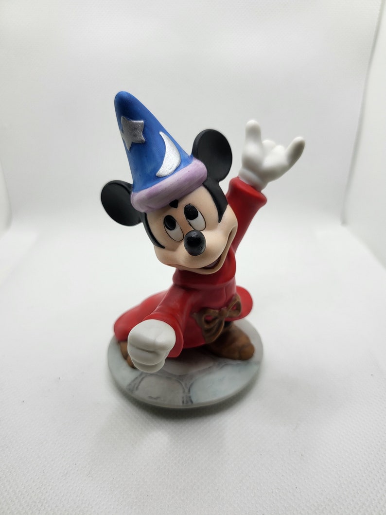 Op de afbeelding: Een keramiek beeldje van Mickey Mouse verkleed als tovenaar. Hij draagt een rode mantel met een blauwe en paarse hoed en houdt zijn hand in de lucht. Hij staat op een ronde basis.
