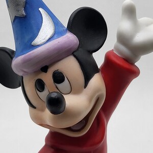 Op de afbeelding: Een keramiek beeldje van Mickey Mouse verkleed als tovenaar. Hij draagt een blauwe hoed met een zilveren halve maan en ster, een rode mantel en zwarte oren. Hij heeft een ondeugende uitdrukking op zijn gezicht.