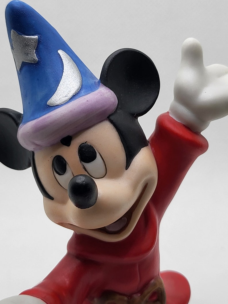 Op de afbeelding: Een keramiek beeldje van Mickey Mouse verkleed als tovenaar. Hij draagt een blauwe hoed met een zilveren halve maan en ster, een rode mantel en zwarte oren. Hij heeft een ondeugende uitdrukking op zijn gezicht.