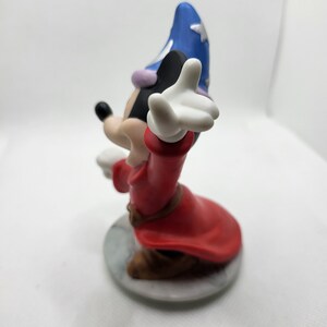 Op de afbeelding: Een keramiek beeldje van Mickey Mouse verkleed als tovenaar. Hij draagt een rode mantel, een blauwe spitse hoed en een witte handschoen. Hij staat op een witte voet.