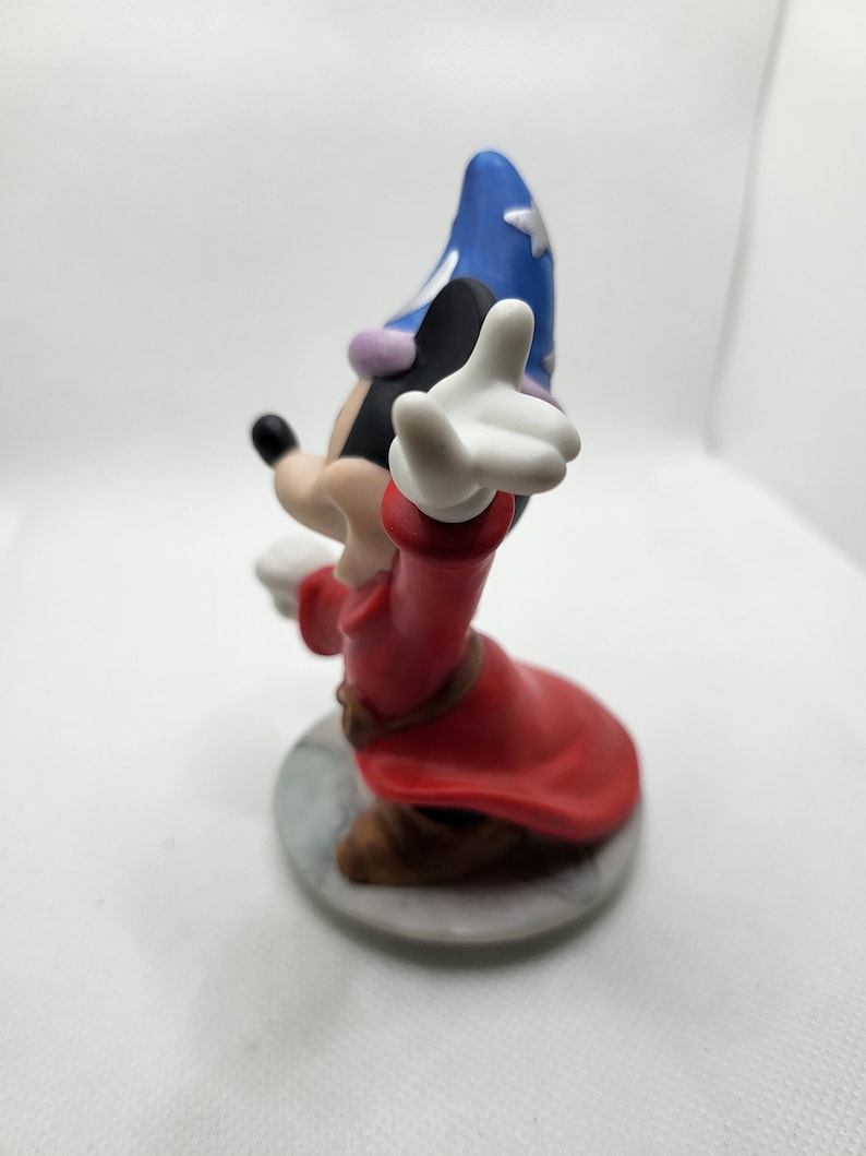Op de afbeelding: Een keramiek beeldje van Mickey Mouse verkleed als tovenaar. Hij draagt een rode mantel, een blauwe spitse hoed en een witte handschoen. Hij staat op een witte voet.