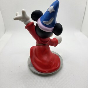 Op de afbeelding: Een keramiek beeldje van Mickey Mouse verkleed als tovenaar. Hij draagt een puntige hoed met een ster en een halve maan, een rode mantel en witte handschoenen. Hij staat op een ronde basis met een grijs en wit marmerpatroon.