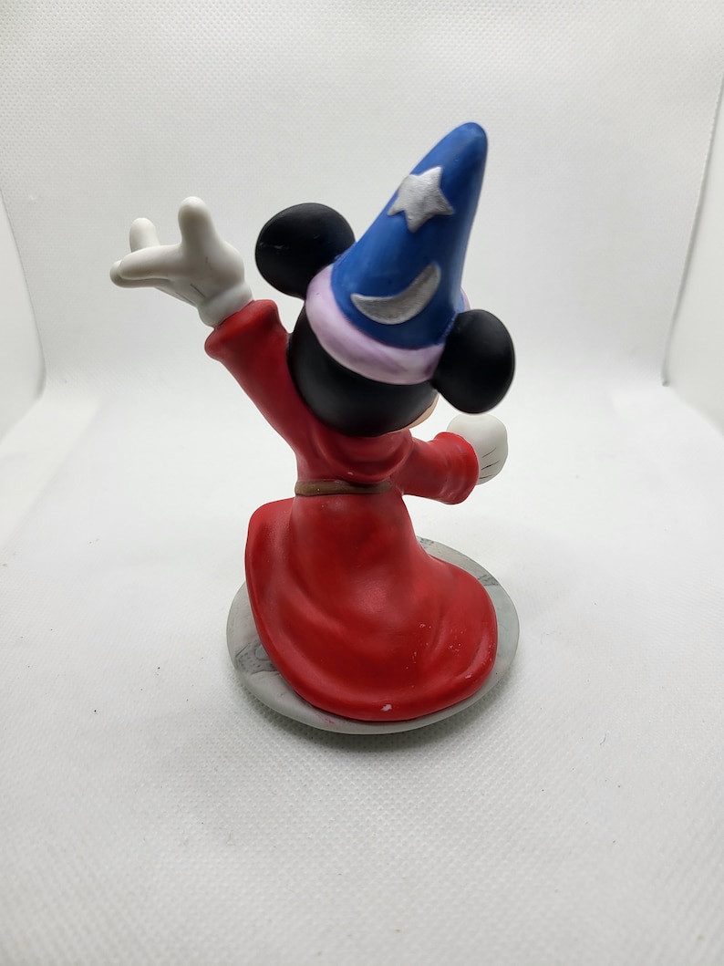 Op de afbeelding: Een keramiek beeldje van Mickey Mouse verkleed als tovenaar. Hij draagt een puntige hoed met een ster en een halve maan, een rode mantel en witte handschoenen. Hij staat op een ronde basis met een grijs en wit marmerpatroon.