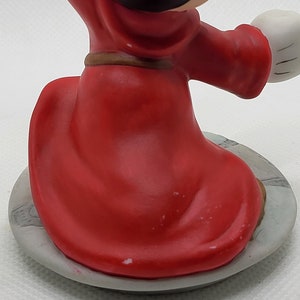 Op de afbeelding: Een rood en zwart keramiek beeldje van Mickey Mouse verkleed als tovenaar. Hij draagt een rode mantel met een bruine riem en een zwarte hoed. Hij staat op een grijze basis.