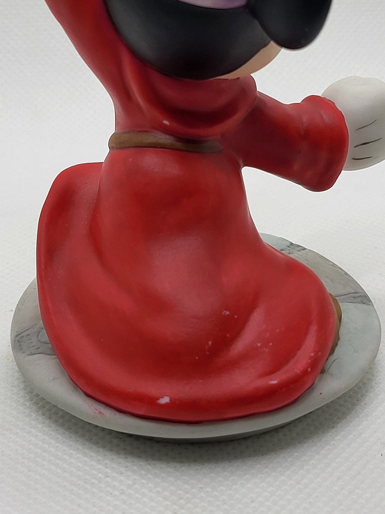Op de afbeelding: Een rood en zwart keramiek beeldje van Mickey Mouse verkleed als tovenaar. Hij draagt een rode mantel met een bruine riem en een zwarte hoed. Hij staat op een grijze basis.