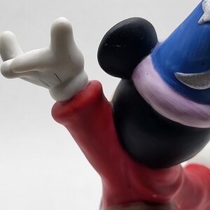 Op de afbeelding: Een keramiek beeldje van Mickey Mouse verkleed als tovenaar. Hij draagt een rode mantel met een bruine riem, een blauwe hoed met een zilveren ster en witte handschoenen. Hij houdt zijn rechterhand omhoog in de lucht, alsof hij een spreuk uitspreekt.