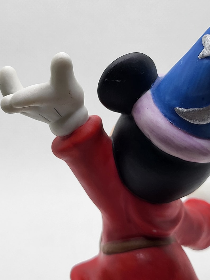 Op de afbeelding: Een keramiek beeldje van Mickey Mouse verkleed als tovenaar. Hij draagt een rode mantel met een bruine riem, een blauwe hoed met een zilveren ster en witte handschoenen. Hij houdt zijn rechterhand omhoog in de lucht, alsof hij een spreuk uitspreekt.