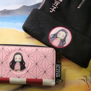 May include: A pink wallet with a black zipper and a Demon Slayer logo. The wallet features a pink and white checkered pattern and a graphic of a character from the anime Demon Slayer. The wallet also includes a black beanie with a pink embroidered character and the Japanese text "鬼滅の刃" (Demon Slayer).