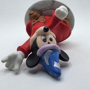 Op de afbeelding: Een keramiek beeldje van Mickey Mouse verkleed als tovenaar. Hij draagt een rode mantel met een blauwe en zilveren puntmuts. Hij houdt een toverstaf in zijn rechterhand en heeft een ondeugende uitdrukking op zijn gezicht.