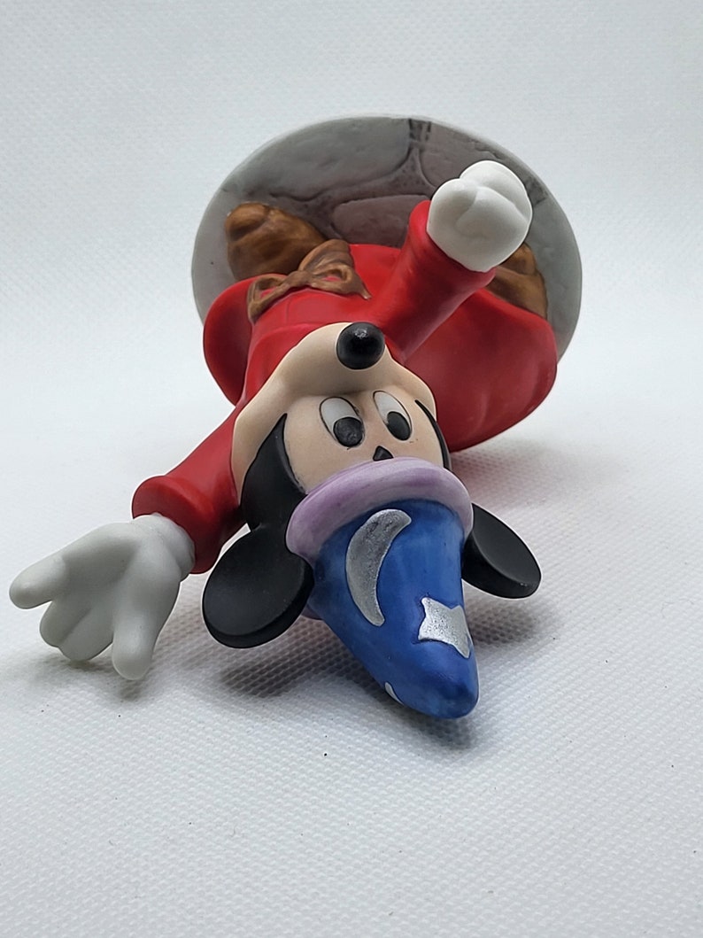 Op de afbeelding: Een keramiek beeldje van Mickey Mouse verkleed als tovenaar. Hij draagt een rode mantel met een blauwe en zilveren puntmuts. Hij houdt een toverstaf in zijn rechterhand en heeft een ondeugende uitdrukking op zijn gezicht.