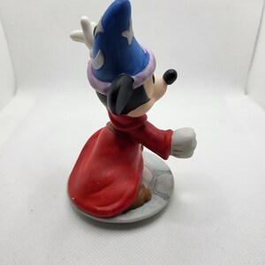 Op de afbeelding: Een keramiek beeldje van Mickey Mouse verkleed als tovenaar. Hij draagt een blauwe en paarse hoed met een ster erop, een rode mantel en witte handschoenen. Hij houdt een staf in zijn rechterhand en knielt.