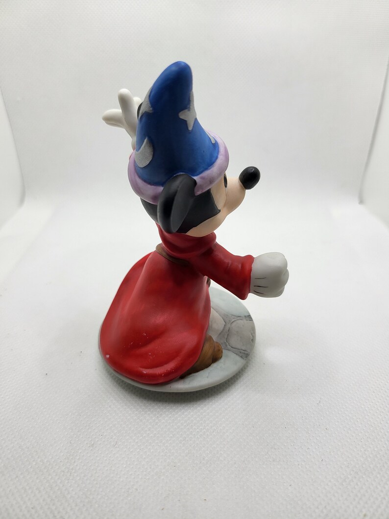 Op de afbeelding: Een keramiek beeldje van Mickey Mouse verkleed als tovenaar. Hij draagt een blauwe en paarse hoed met een ster erop, een rode mantel en witte handschoenen. Hij houdt een staf in zijn rechterhand en knielt.