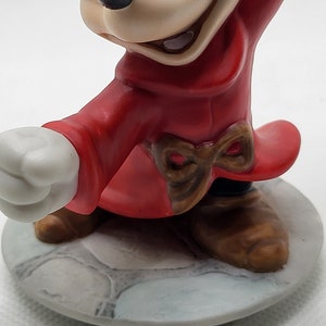 Op de afbeelding: Een keramiek beeldje van Mickey Mouse verkleed als tovenaar. Hij draagt een rode mantel met een bruine strik en bruine schoenen. Hij staat op een grijze stenen basis.