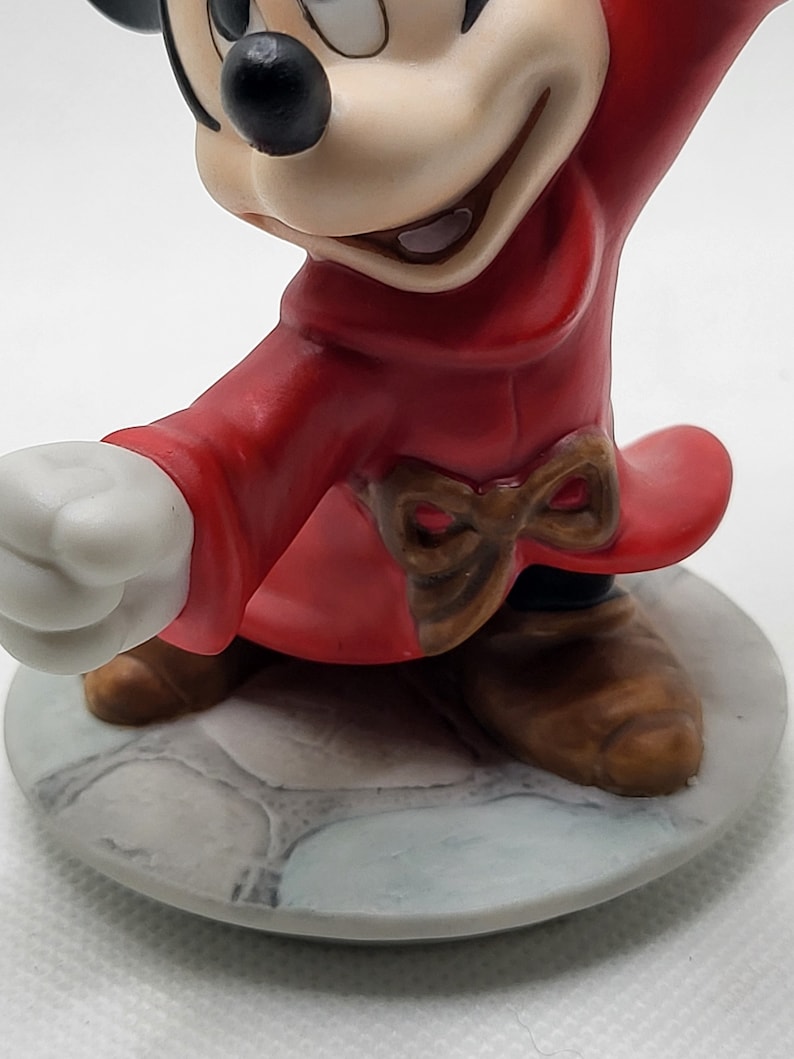 Op de afbeelding: Een keramiek beeldje van Mickey Mouse verkleed als tovenaar. Hij draagt een rode mantel met een bruine strik en bruine schoenen. Hij staat op een grijze stenen basis.