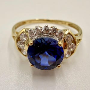 May include: A gold ring featuring a large, round, deep blue center stone held by four prongs. The setting is accented with a cluster of small, clear, round-cut stones arranged in a semi-circle above and on both sides of the central blue stone.