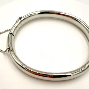 May include: A shiny, silver-colored hinged bangle bracelet. The bracelet is a complete circle with a clasp and a small chain for security. The metal reflects light, creating highlights and shadows. The bracelet is on a white surface.