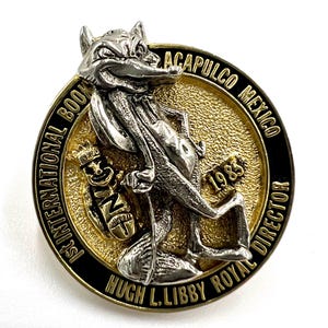 May include: A vintage pin with a silver fox-like character holding a staff, set against a gold-toned background. The pin is inscribed with "ACAPULCO MEXICO 1983" and "1st INTERNATIONAL BOOK DIRECTOR".