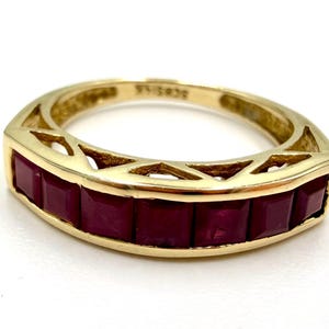 May include: A gold ring with a row of rectangular, deep red gemstones. The ring has a geometric design with cut-out shapes along the band. The gemstones are set in a channel setting, creating a continuous line of color.
