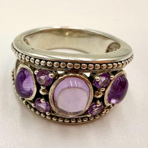 May include: A wide silver band ring featuring a central large oval purple amethyst cabochon, flanked by two smaller oval amethyst cabochons, and accented with four small round faceted purple amethyst stones and beaded metal detailing.