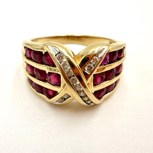 May include: A gold ring featuring three horizontal rows of round, deep red rubies set within a band. The center of the ring displays a prominent, crossed X-shaped design embellished with a single row of small, clear, sparkling white diamonds.