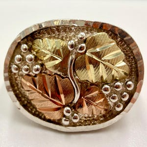 May include: Oval-shaped silver-tone ring with a gold and rose gold leaf design. The leaves are detailed with veins and surrounded by small silver-tone beads. The ring has a textured silver border.