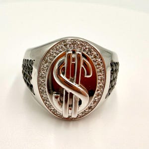 May include: A silver-toned ring featuring a dollar sign emblem. The ring has a central oval design with a silver dollar sign, surrounded by small, clear stones. The sides of the ring have a black, textured design.