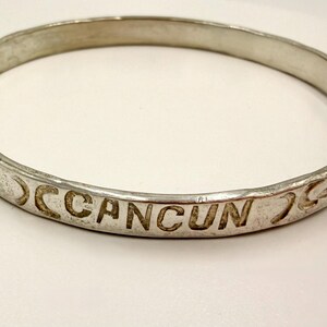 May include: A silver-toned metal bangle bracelet featuring a smooth, rounded band. The exterior surface is engraved with the word CANCUN in capital letters, flanked by stylized eye motifs on both sides of the text, creating a continuous pattern.