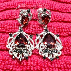 May include: A pair of silver-toned drop earrings with large, teardrop-shaped, dark red gemstones. The earrings have an ornate, filigree design with smaller black accents and a post back. The background is a bright pink, textured fabric.