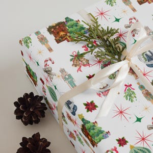May include: A white gift box wrapped in paper featuring a repeating pattern of nutcrackers, candy canes, Christmas trees, poinsettias, bells, and the word JOY in red letters, tied with a cream satin ribbon and topped with a green sprig.