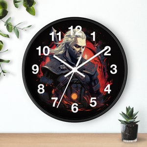 Op de afbeelding: Zwarte wandklok met een witte wijzerplaat en witte wijzers. De wijzerplaat van de klok heeft een afbeelding van een personage uit het videospel The Witcher. De cijfers op de wijzerplaat van de klok zijn wit.