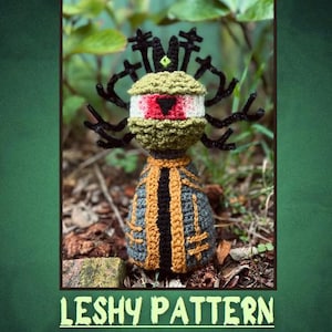 May include: A crocheted Leshy doll featuring a round olive green head with a single large eye, a black triangular pupil, and a crown of black antler-like branches. The body is a grey cone shape with gold and black vertical trim. Text reads LESHY PATTERN.
