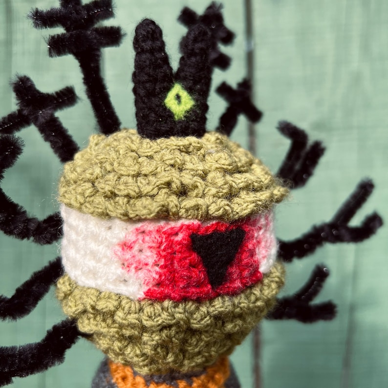 May include: A crocheted figure featuring a textured olive green head with a white band across the center, accented by a red patch and a black triangle. A black crown with a lime green eye sits on top, surrounded by black pipe cleaner branches.