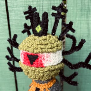 May include: A crocheted figure featuring a round olive green head with a white band across the center, a red patch, and a black triangular detail. It has a black crown with a lime green accent and black pipe cleaner branches extending outward.