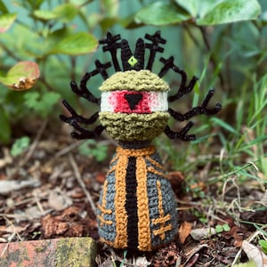 May include: A handmade crochet doll featuring a round olive green head with a large central eye, a black triangular pupil, and a small green eye on top. The body is dark gray with orange and black vertical accents and black branch-like appendages.