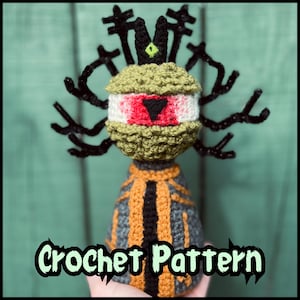 May include: A crocheted figure featuring a large central eye with a red iris and black triangular pupil, topped with a black crown-like structure and branching black antlers. The body is gray with orange vertical stripes and black trim.