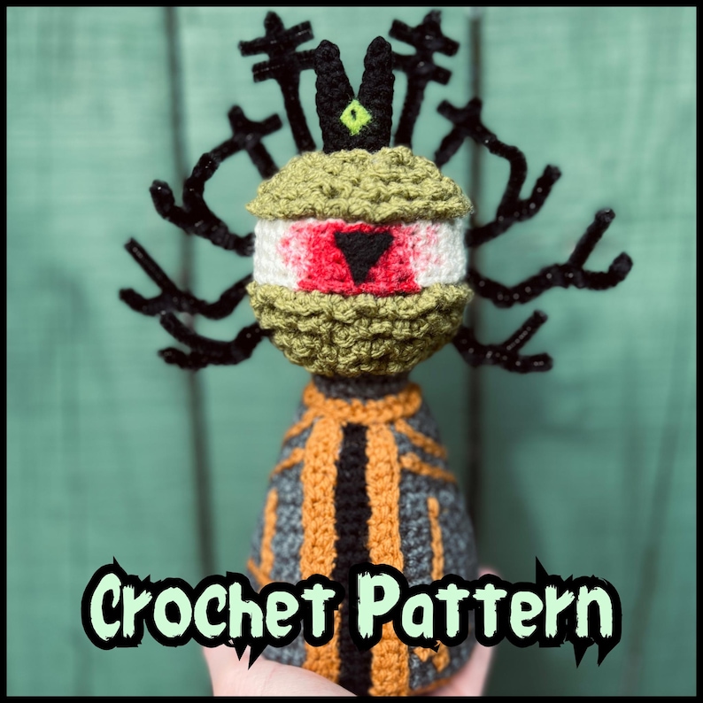 May include: A crocheted figure featuring a large central eye with a red iris and black triangular pupil, topped with a black crown-like structure and branching black antlers. The body is gray with orange vertical stripes and black trim.