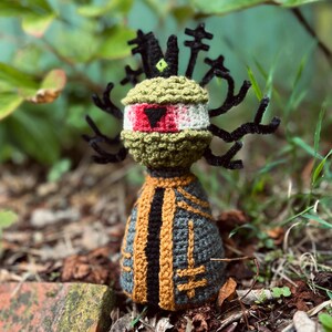 May include: A handmade crochet doll featuring a round olive green head with a large red eye and a black triangular pupil. The doll has black pipe cleaner antennae and wears a grey and gold robe with a central black stripe and cross-like symbols.