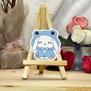 May include: A sticker featuring a white bunny wearing a blue frog costume with a bow tie. The sticker is displayed on a small wooden easel. The background includes roses and a wrapped gift.