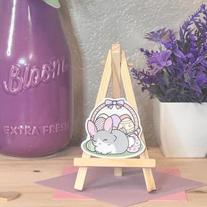 May include: A sticker featuring a gray bunny resting next to a basket of Easter eggs. The sticker is displayed on a small wooden easel. The background includes a purple vase and a pot of purple flowers.