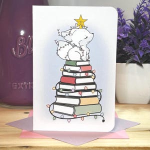 Könnte beinhalten: Eine weiße Grußkarte mit einer skurrilen Illustration. Sie zeigt einen Fuchs, der auf einem Stapel Bücher sitzt, die wie ein Weihnachtsbaum dekoriert sind, verziert mit Lichterketten und einem goldenen Stern. Die Bücher sind in verschiedenen Farben.