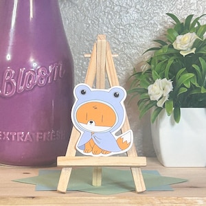 Può includere: Un adesivo con un personaggio di una volpe dei cartoni animati con una felpa con cappuccio a forma di rana blu chiaro. La volpe è arancione e bianca, con una coda bianca. L'adesivo è esposto su un piccolo cavalletto di legno.