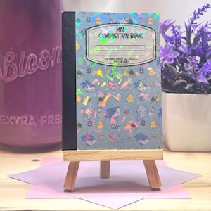 Könnte beinhalten: Ein kleines Notizbuch mit blauem Einband, das ein Muster aus Cartoon-Meereskreaturen und den Text "MES COMPOSITION BOOK" zeigt. Das Buch wird auf einer Miniatur-Holzstaffelei präsentiert.