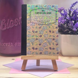 Puede incluir: Mini cuaderno de composición con una cubierta marrón que presenta un patrón de hipopótamos grises y detalles holográficos. El cuaderno se exhibe en un pequeño caballete de madera. El texto "Mini Composition Book" es visible.