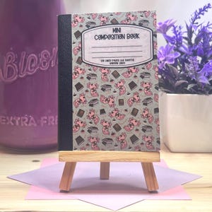 Peut inclure: Un mini cahier de composition avec une couverture grise ornée d'un motif d'axolotls roses et de livres. Le cahier est présenté sur un petit chevalet en bois. Le texte "Mini Composition Book" est visible.