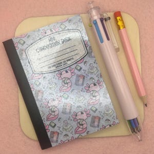 Peut inclure: Un mini cahier de composition avec une couverture grise ornée d'illustrations d'axolotls, un stylo multicolore et un crayon rose avec une gomme. Le cahier porte l'inscription "Mini Composition Book" et "128 Lined Pages (64 Sheets) Narrow Lined".