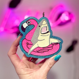 May include: A glittery ornament featuring a pink flamingo float with a shark biting into it. The ornament has a blue border and a silver loop for hanging. The flamingo is pink, and the shark is gray and yellow.