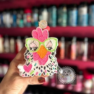 May include: A whimsical, handmade chicken-shaped ornament. The ornament features a pink comb, an orange beak, and green-framed glasses. The chicken's body is white with black and colorful polka dots, and a pink heart. A sparkly ball and a red bead are at the top.
