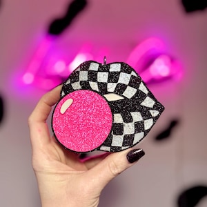 May include: A decorative ornament shaped like a pair of lips blowing a bubble. The lips have a black and white checkered pattern with silver glitter accents. The bubble is a bright pink color.