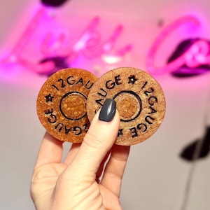 May include: Two circular, amber-colored resin coasters with a textured, glittery finish. Each piece features the embossed black text 12 GAUGE and a star symbol, mimicking the base of a shotgun shell. The design includes a central circular indentation.